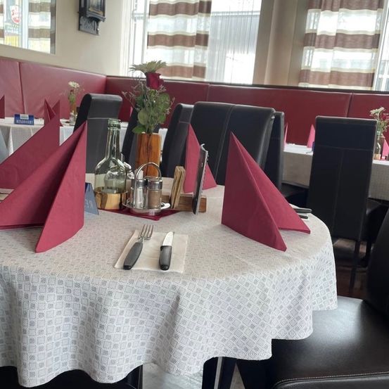 Ein Restauranttisch mit roten Servietten, Besteck und einer Blumenvase. Es gibt Stühle um den Tisch herum. Im Hintergrund befinden sich weitere Tische und ein Fenster mit Vorhängen.