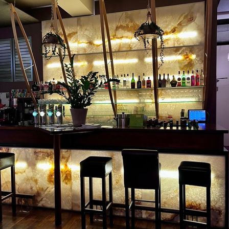 Eine Bartheke mit modernem Design, ausgestattet mit schwarzen Stühlen, einer großen Topfpflanze und einer Marmorrückwand. Verschiedene Flaschen und Gläser sind auf den Regalen ausgestellt, und ein Monitor ist auf der Theke platziert.