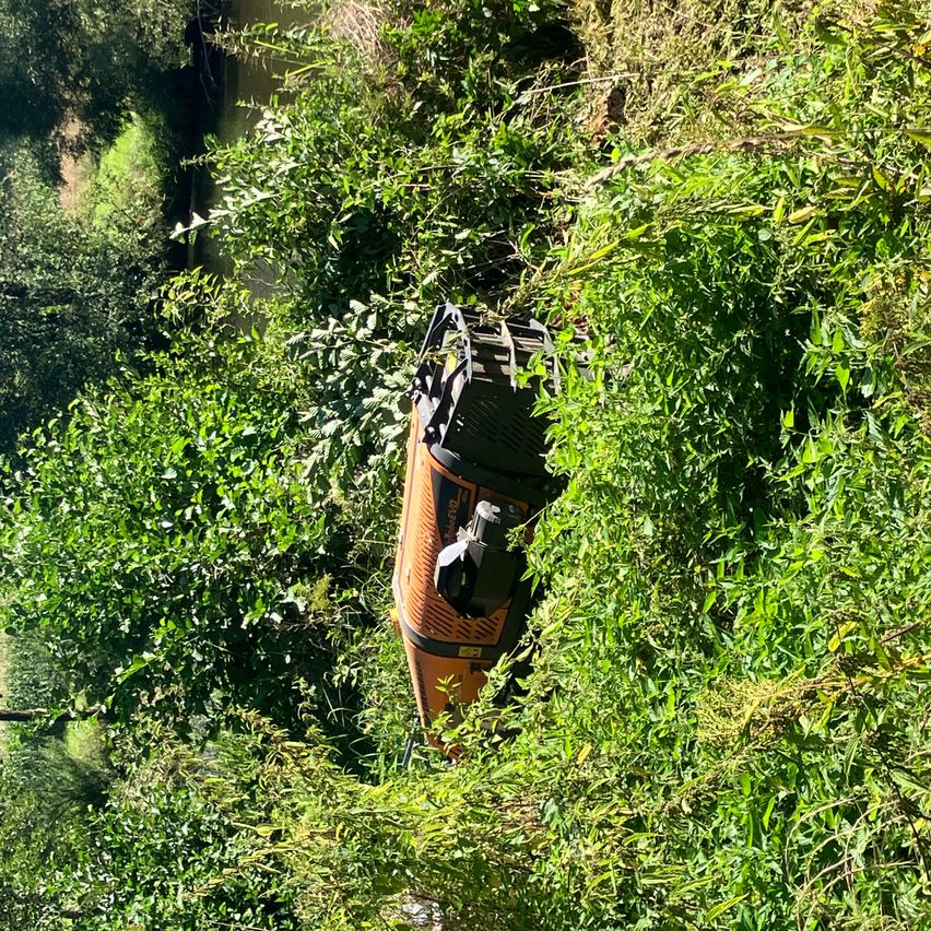 Eine Vogelperspektive einer orangefarbenen und schwarzen Maschine, die in einem Wald feststeckt, umgeben von hohen, üppigen grünen Pflanzen.