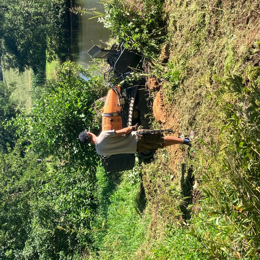 Ein Mann benutzt eine große orangefarbene Maschine, um den Boden zu räumen, und steht auf einem Hang in der Nähe eines Flusses mit üppiger Vegetation.