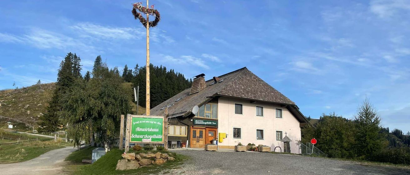 Ein weißes Haus mit einem braunen Dach steht in einer Lichtung. Ein grünes Schild mit den Worten Almiwirtshaus Schwarzarhogehalle steht davor. Bäume und ein Pfahl mit einer herzförmigen Dekoration sind in der Nähe.