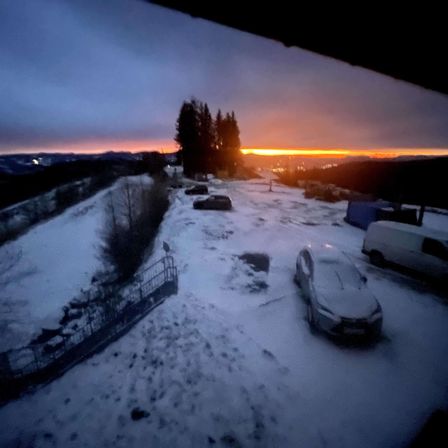Eine verschneite Landschaft mit einem Sonnenuntergang im Hintergrund. Autos sind am Straßenrand geparkt, und ein Van ist rechts geparkt. Der Himmel ist orange und pink, und es gibt Bäume in der Ferne.