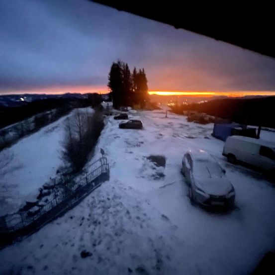 Eine verschneite Landschaft mit einem Sonnenuntergang im Hintergrund. Autos sind am Straßenrand geparkt, und ein Van ist rechts geparkt. Der Himmel ist orange und pink, und es gibt Bäume in der Ferne.