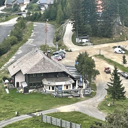 Ein Luftbild eines Chalets, umgeben von Bäumen, mit mehreren geparkten Autos in der Nähe. Ein Pickup-Truck ist auf der unbefestigten Straße, und ein weißer Lieferwagen ist am Straßenrand geparkt. Es gibt eine grasbewachsene Wiese und einen Betonweg, der zum Chalet führt.
