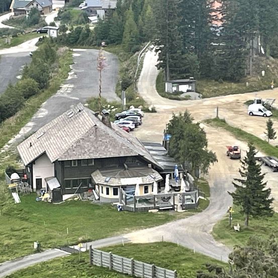 Ein Luftbild eines Chalets, umgeben von Bäumen, mit mehreren geparkten Autos in der Nähe. Ein Pickup-Truck ist auf der unbefestigten Straße, und ein weißer Lieferwagen ist am Straßenrand geparkt. Es gibt eine grasbewachsene Wiese und einen Betonweg, der zum Chalet führt.