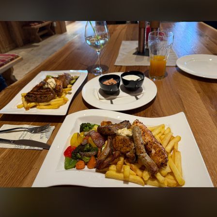 Zwei Teller mit Essen stehen auf einem Holztisch. Ein Teller hat ein Steak, Pommes Frites und zwei Saucen. Der andere hat Hähnchen, Würste, Pommes Frites und Gemüse.