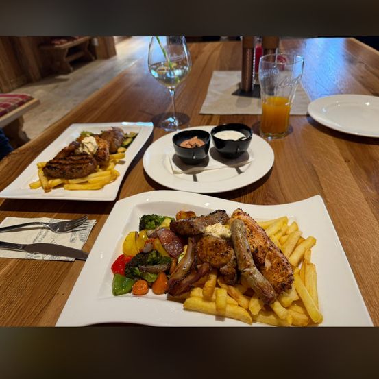 Zwei Teller mit Essen stehen auf einem Holztisch. Ein Teller hat ein Steak, Pommes Frites und zwei Saucen. Der andere hat Hähnchen, Würste, Pommes Frites und Gemüse.