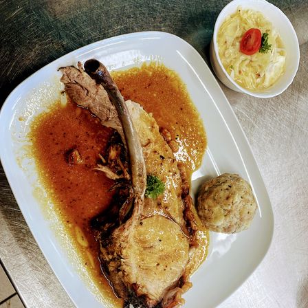 Ein Teller mit gebratenem Schweinebraten in Soße und einer Beilage von Knödeln und Sauerkraut. Der Braten ist mit Kräutern garniert.