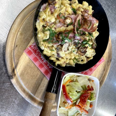 Eine Pfanne mit cremiger Mac and Cheese mit Speck, Zwiebeln und Champignons, garniert mit Petersilie, serviert auf einem Holzbrett mit einem Beilagensalat.