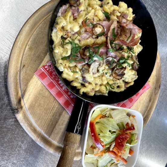 Eine Pfanne mit cremiger Mac and Cheese mit Speck, Zwiebeln und Champignons, garniert mit Petersilie, serviert auf einem Holzbrett mit einem Beilagensalat.