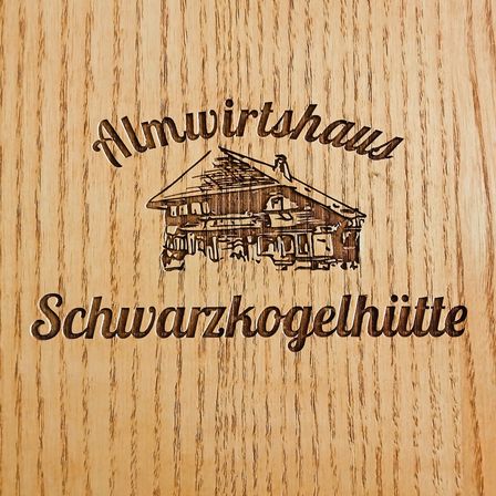 Eine hölzerne Oberfläche mit einem Logo von Almwirtshaus Schwarzkogelhütte. Das Logo zeigt ein Haus mit Schornstein.