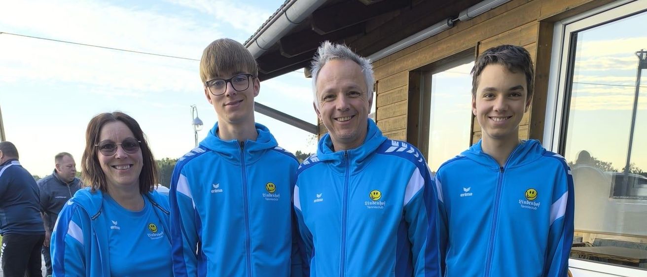 Vier Personen in blauen Jacken stehen lächelnd zusammen. Zwei Männer und eine Frau mit einem lächelnden Jungen neben ihnen. Dahinter ein Holzgebäude und Menschen in der Ferne.