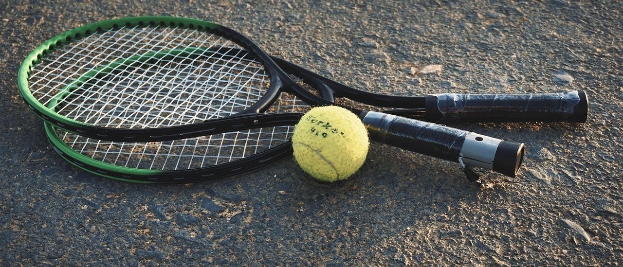 Zwei Tennisschläger liegen auf einem Platz mit einem Tennisball dazwischen. Die Schläger sind schwarz mit grünen Akzenten, und der Ball ist gelb mit dem Wort 'Wilson' darauf geschrieben.