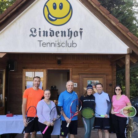 Eine Gruppe von Menschen steht vor einem hölzernen Gebäude mit einem lächelnden Gesichtslogo und der Aufschrift 'Lindenhof Tennisclub'. Sie halten alle Tennisschläger.