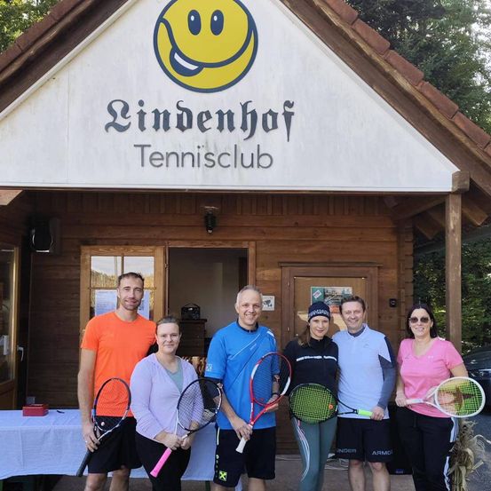Eine Gruppe von Menschen steht vor einem hölzernen Gebäude mit einem lächelnden Gesichtslogo und der Aufschrift 'Lindenhof Tennisclub'. Sie halten alle Tennisschläger.