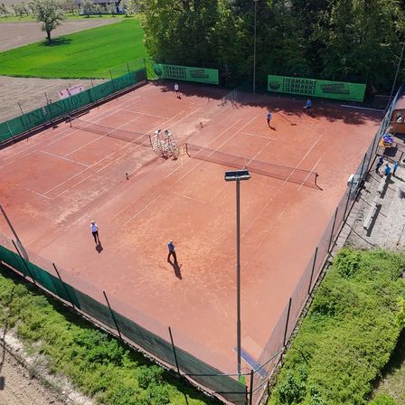 Eine Luftaufnahme eines Tennisplatzes mit mehreren verstreuten Spielern. Der Platz hat grüne und rote Oberflächen, mit grünen Zäunen rundherum. Werbetafeln sind an den Zäunen montiert.
