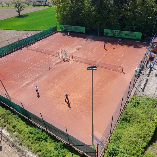 Eine Luftaufnahme eines Tennisplatzes mit mehreren verstreuten Spielern. Der Platz hat grüne und rote Oberflächen, mit grünen Zäunen rundherum. Werbetafeln sind an den Zäunen montiert.