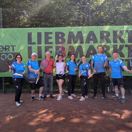 Eine Gruppe von Tennisspielern in blauen Uniformen steht auf einem Tennisplatz und hält Tennisschläger in der Hand. Hinter ihnen zeigt eine grüne Banner die Textur 'Liebemarkt' an.