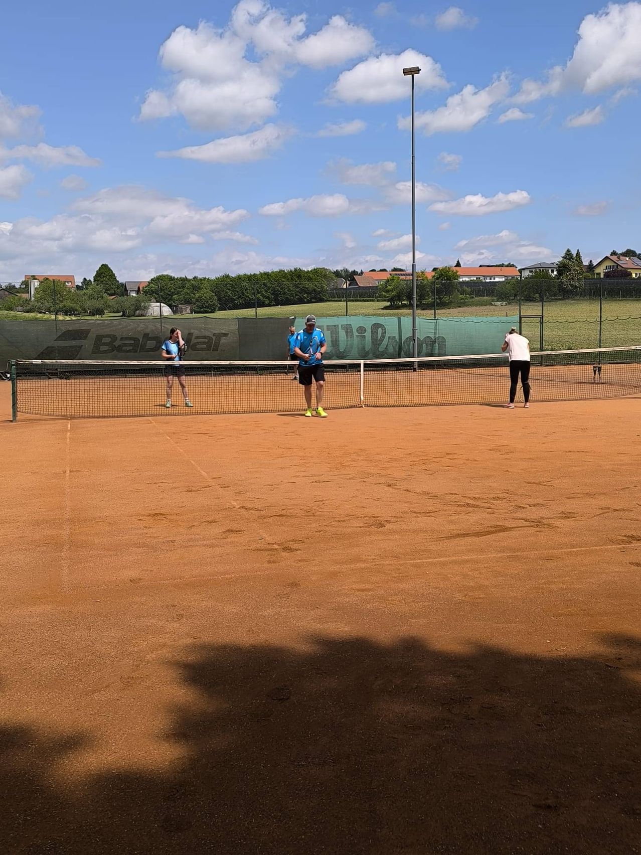 Tennisspieler bereiten sich auf ein Match auf einem Sandplatz mit Netz, Zaun und Häusern in der Ferne vor. Ein Spieler hält einen Schläger.