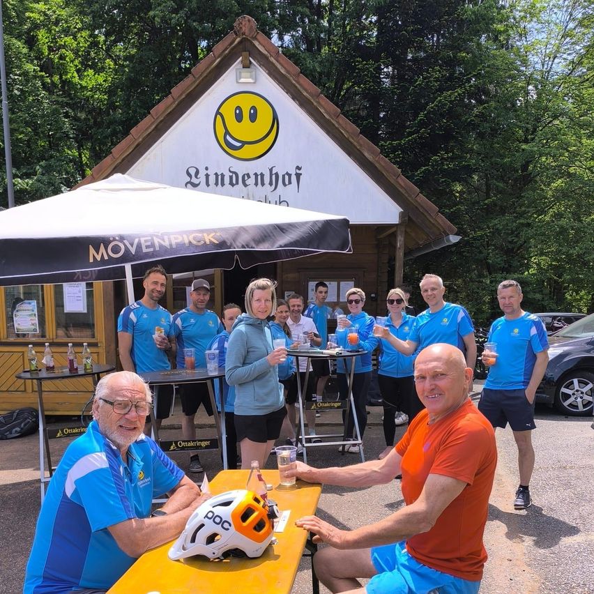 Eine Gruppe von Radfahrern posiert für ein Foto vor einem Gebäude mit einem lächelnden Gesichtslogo. Sie sind in blauen und weißen Uniformen gekleidet.