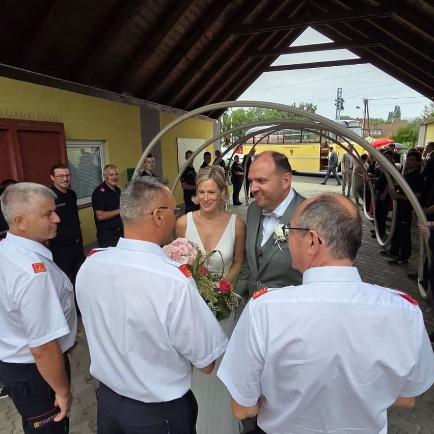 Eine Hochzeitszeremonie in einem Unterstand, mit einem Bräutigam und einer Braut, umgeben von Leuten in Uniformen, und einem gelben Bus im Hintergrund.