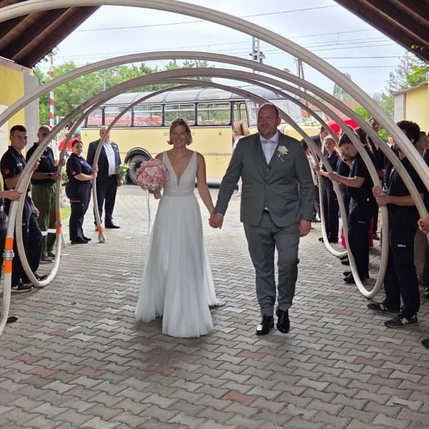 Eine Braut und ein Bräutigam gehen Hand in Hand durch einen Bogen, während eine Menge hinter ihnen jubelt. Die Braut hält einen Blumenstrauß und der Bräutigam trägt einen Anzug und Lederschuhe.