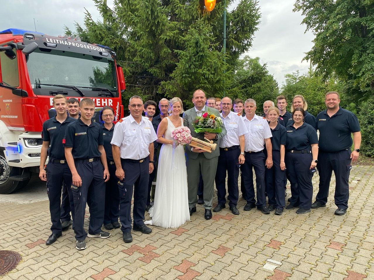 Ein Hochzeitspaar steht mit einem Blumenstrauß und einer Trophäe, umgeben von Feuerwehrleuten in Uniform. Sie posieren vor einem Feuerwehrwagen.