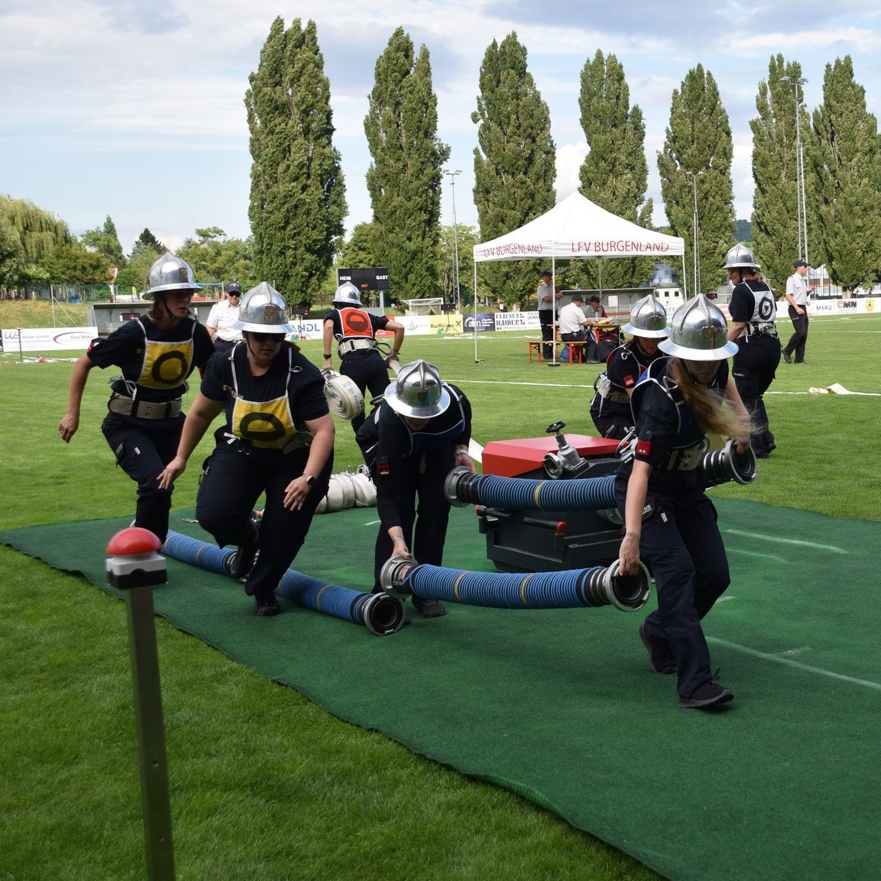 Eine Gruppe von Feuerwehrleuten trainiert auf einem Feld. Sie tragen Helme und Uniformen und arbeiten mit einer Feuerwehrhose. Ein Zelt ist im Hintergrund.