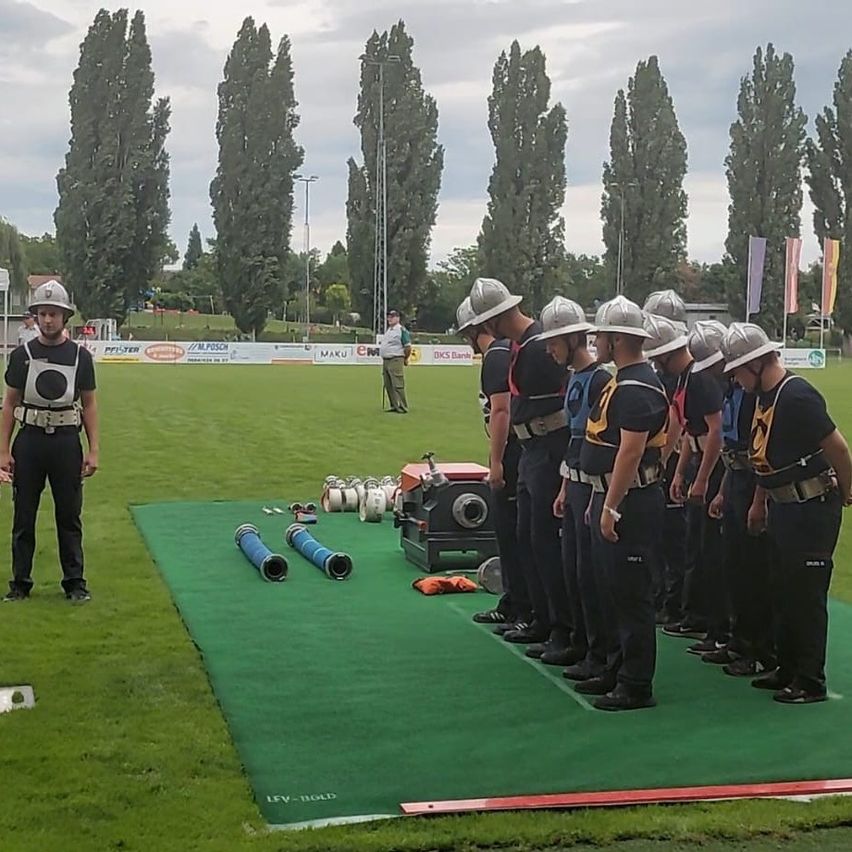 Eine Gruppe von Personen in Schutzkleidung und Helmen steht auf einem grünen Feld, wahrscheinlich in Vorbereitung auf eine Veranstaltung. Eine Person vorne trägt eine weiße Schürze. Mehrere Gegenstände, darunter Schläuche und Fässer, sind auf dem Feld platziert.
