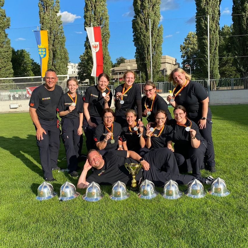 Eine Gruppe von Menschen posiert für ein Foto mit Medaillen und Trophäen auf einem Rasenfeld. Dahinter befinden sich Fahnen und Gebäude.