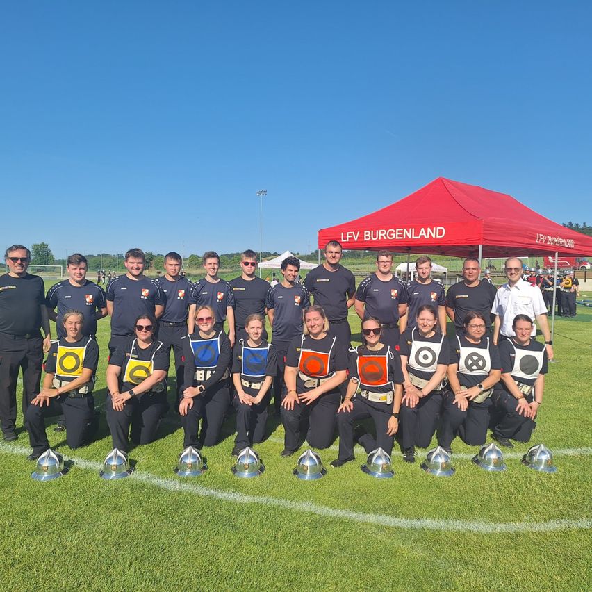 Eine Gruppe von Menschen in Uniform posiert für ein Foto auf einem Feld mit Trophäen vor ihnen. Sie befinden sich in der Nähe eines Zeltes mit den Worten 'LFV Burgenland'.