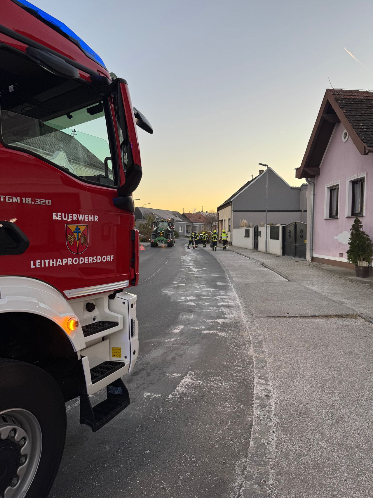 A fire truck from Leithaprodersdorf is parked on the side of the road. Several firefighters are walking on the street. A building with a pink facade and a green plant in front is visible.