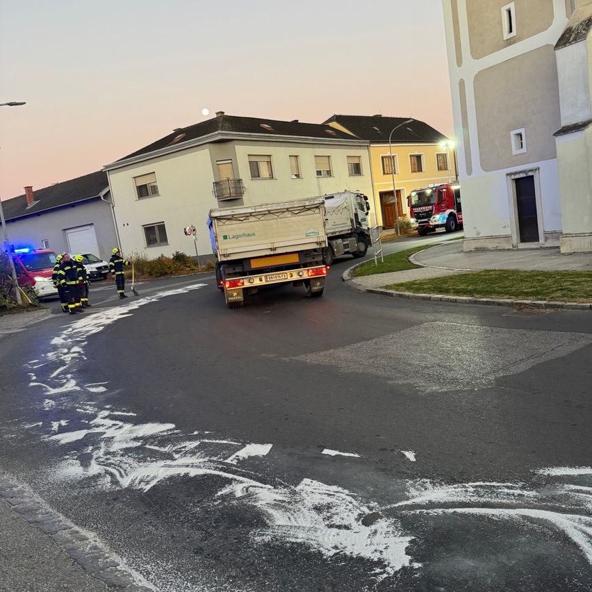 Emergency vehicles are parked near a road with white markings, and firefighters are standing on the sidewalk. Buildings line the street.