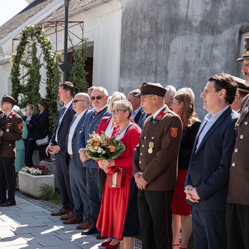 A group of formally dressed people stand in line outside. A woman in a red dress holds a bouquet of flowers. Behind them, a building with plants is visible.