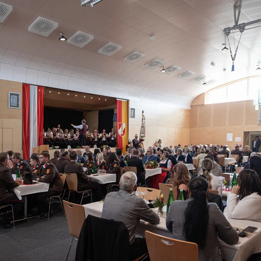 Ein großer Saal mit Tischen und Stühlen, gefüllt mit Menschen, die an einer Veranstaltung teilnehmen. Eine Band spielt auf der Bühne mit Fahnen auf beiden Seiten. Das Publikum sitzt und schaut zu.