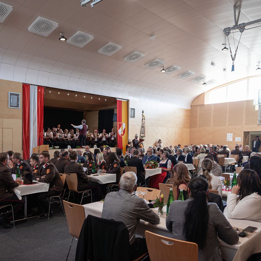 A large hall with tables and chairs filled with people attending an event. A band is performing on the stage with flags on either side. The audience is seated and watching.