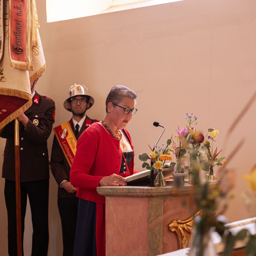 A woman speaks into a microphone at a podium, flowers and flags behind her. Two men stand behind her, one in a firefighter uniform.
