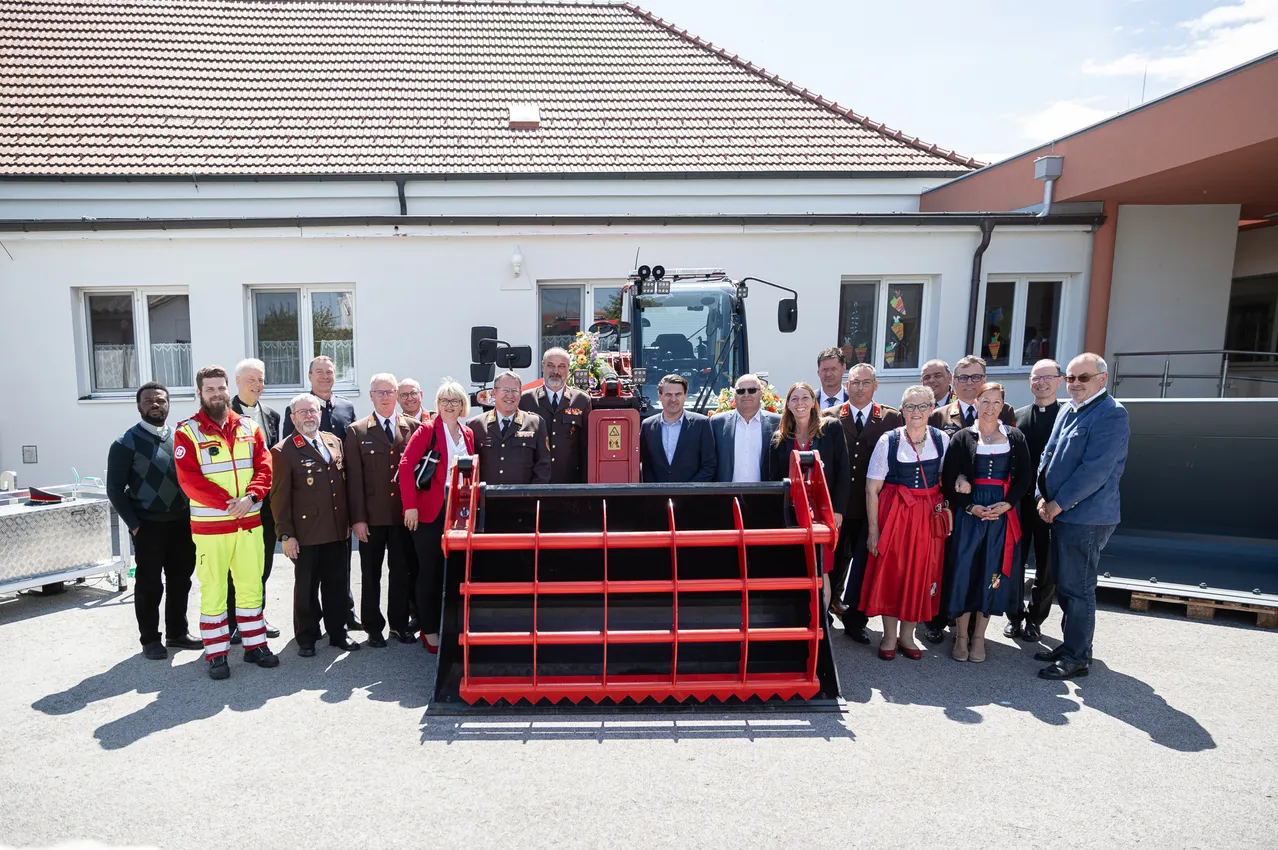 Eine Gruppe von Menschen ist vor einem Gebäude mit einem roten Maschinenfahrzeug versammelt. Sie sind alle in formeller Kleidung und einige tragen Uniformen. Sie alle lächeln und posieren für ein Foto.
