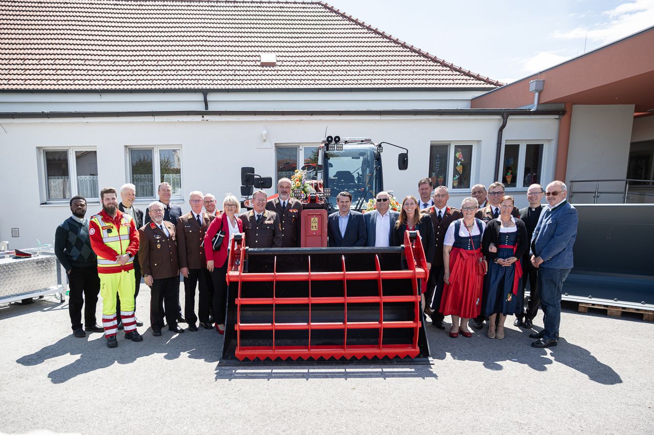 A group of people are gathered in front of a building with a red machinery vehicle. They are all dressed in formal attire, and some are wearing uniforms. They are all smiling and posing for a photo.