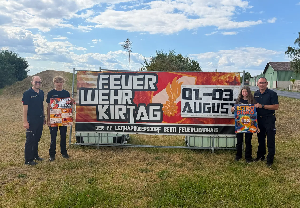 Zwei Personen stehen vor einem großen Banner mit den Worten 'Feuer Wehr Kirtag' und '01-03 August'. Sie halten Werbematerialien in den Händen und scheinen eine Veranstaltung zu bewerben. Das Banner ist vor einer Kulisse aus einem Grasfeld mit Bäumen und einem bewölkten Himmel.