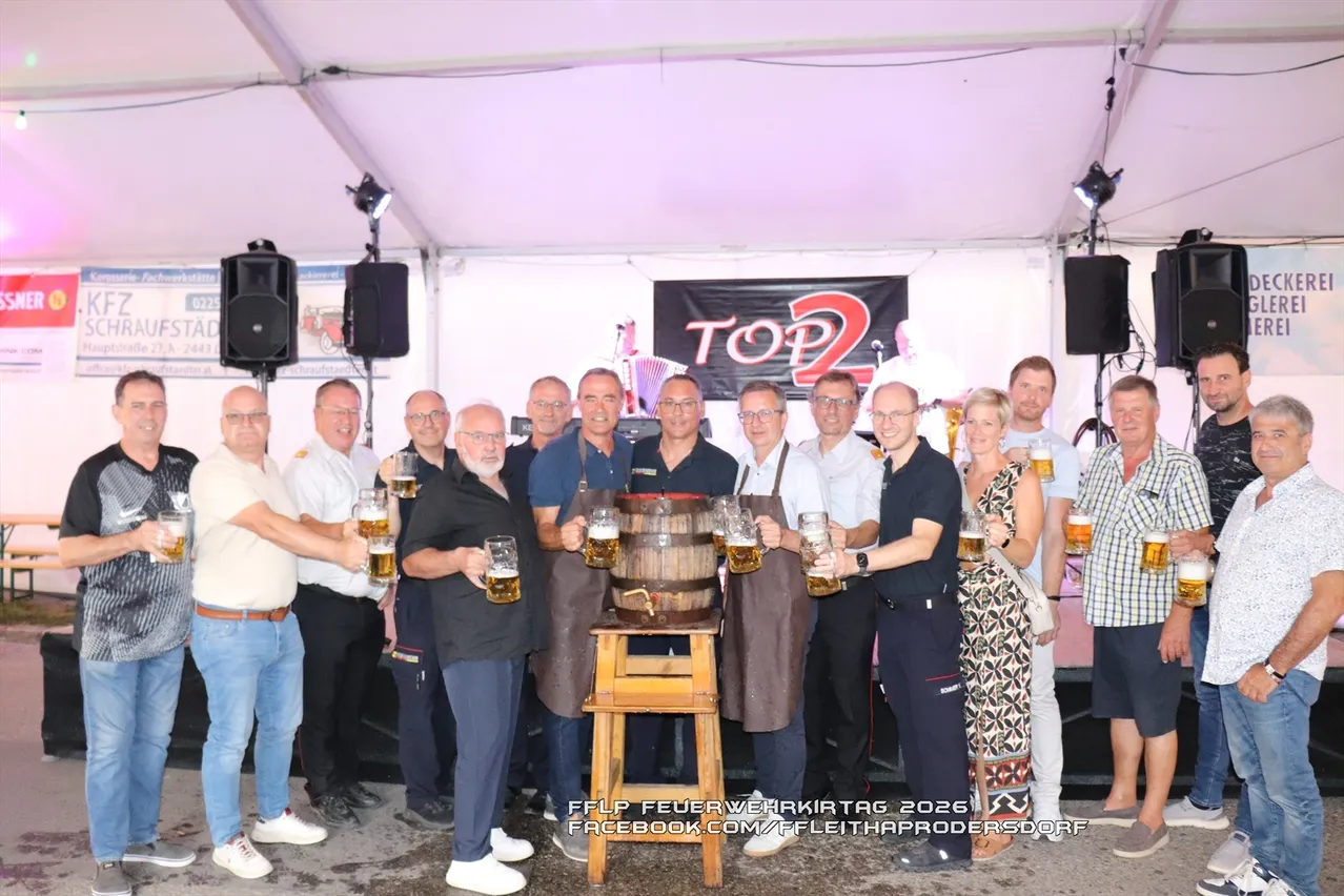 Eine Gruppe von Menschen in einem Zelt, die Biergläser halten, mit einem Fass in der Mitte und einem Banner mit der Aufschrift 'TOP2' im Hintergrund.