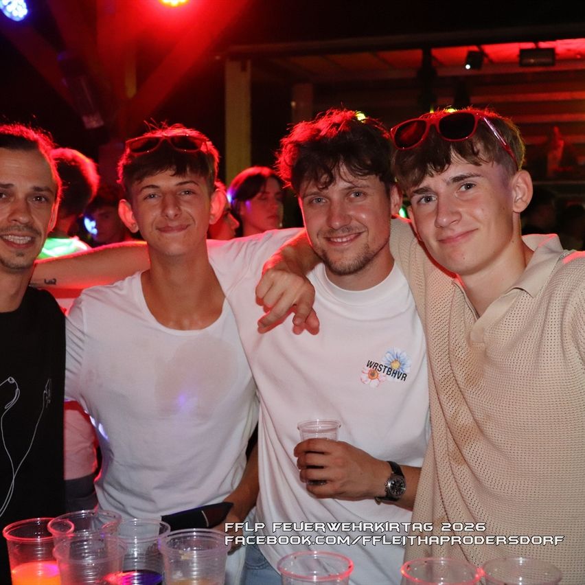 Vier junge Männer posieren für ein Foto in einem Club mit roter Beleuchtung, einer hält ein Getränk. Sie alle lächeln und scheinen sich zu amüsieren.