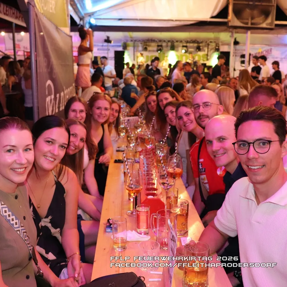 Eine Gruppe von Menschen sitzt an einem langen Tisch mit Gläsern Bier, lächelt und posiert für ein Foto bei einer Outdoor-Veranstaltung.