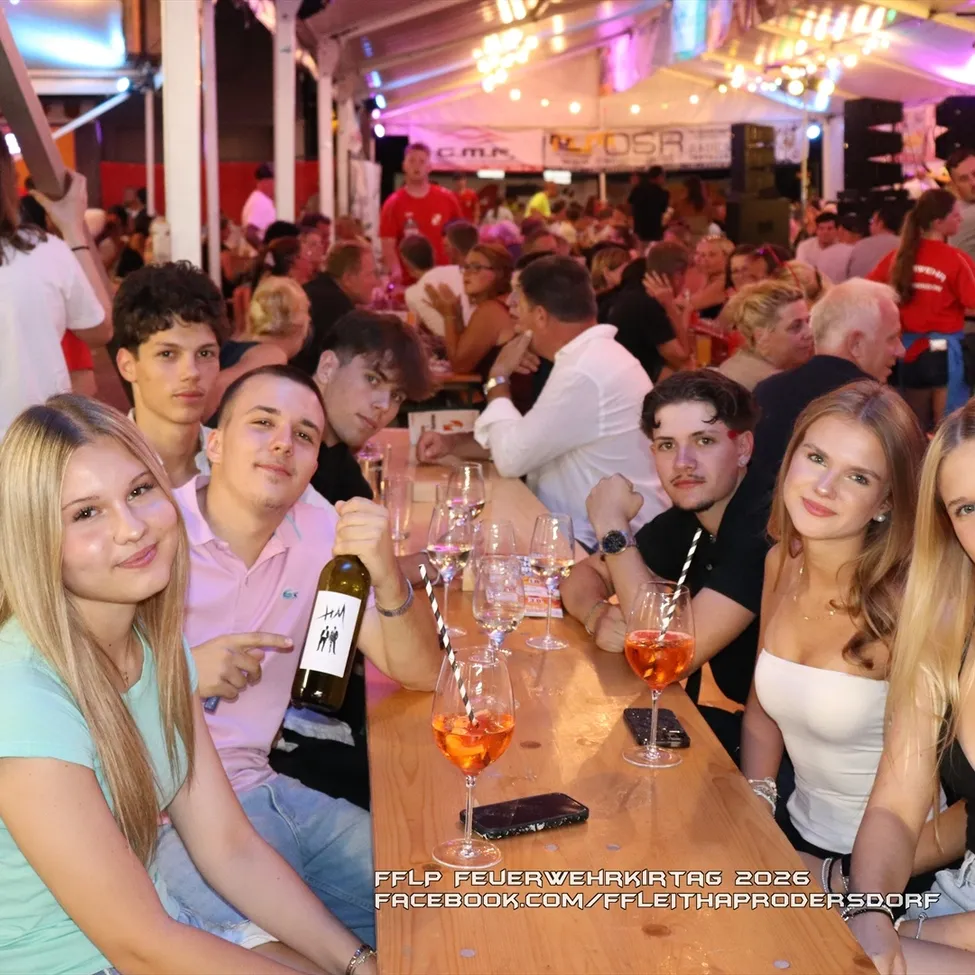 Eine Gruppe junger Erwachsener, sowohl Männer als auch Frauen, sitzen an einem langen Tisch bei einer lebhaften Outdoor-Veranstaltung, lächeln und posieren für ein Foto. Sie haben Weingläser, eine Flasche und Handys auf dem Tisch. Im Hintergrund ist ein überfüllter Bereich mit stehenden und sitzenden Personen zu sehen.