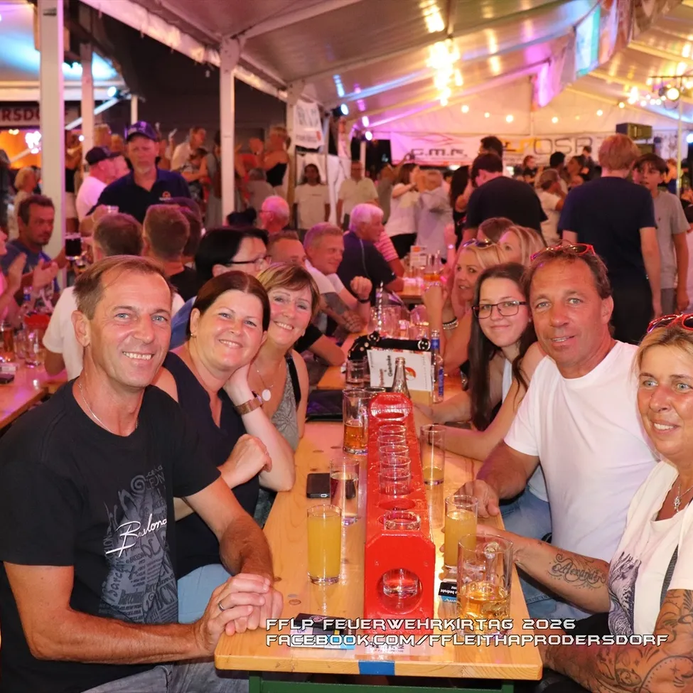 Eine Gruppe von Menschen sitzt an einem Tisch mit Biergläsern und einem roten Bierhahn in der Mitte, lächelt und posiert für ein Foto bei einer Outdoor-Veranstaltung.