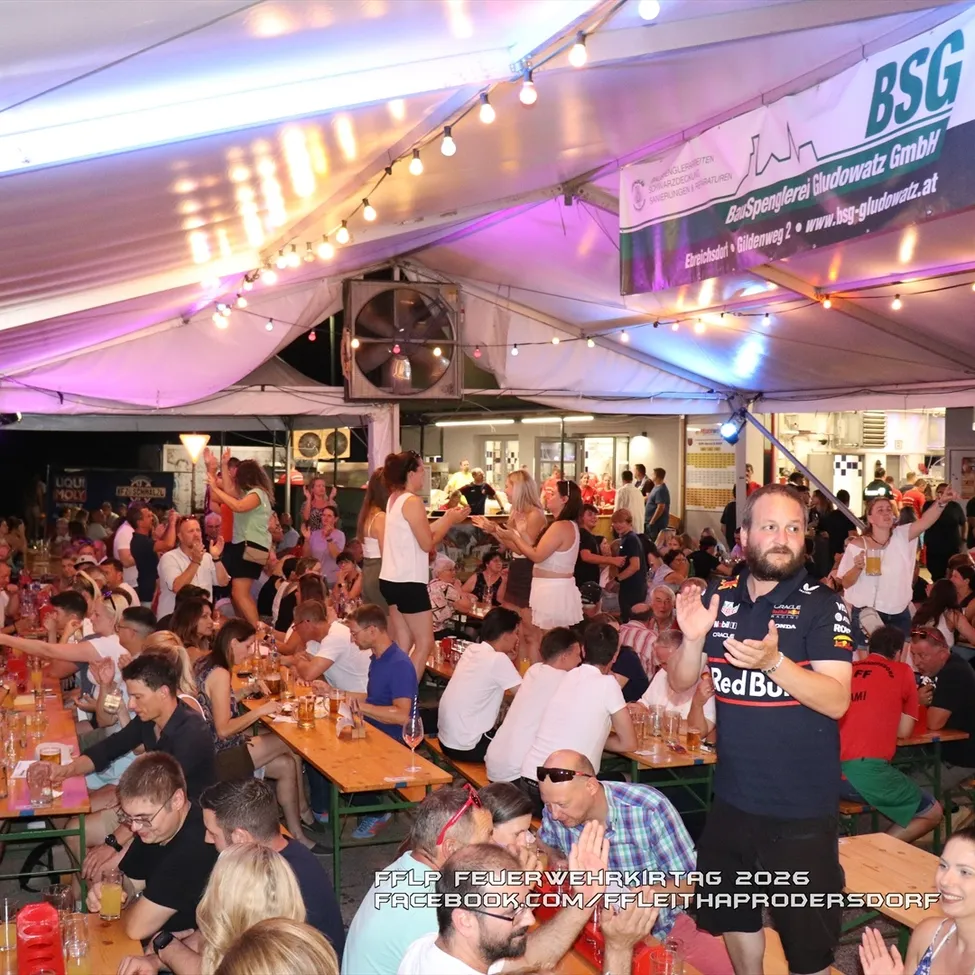 Eine große Menschenmenge versammelt sich bei einer Outdoor-Veranstaltung, möglicherweise einem Festival, unter einem Zelt mit einem Banner, das 'BSG' liest. Viele Menschen sitzen an Tischen, trinken und plaudern, während andere stehen und tanzen. Die Atmosphäre ist lebhaft und festlich.