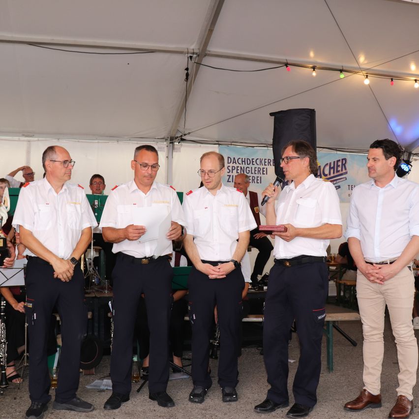 Fünf Männer in Uniform stehen unter einem Zelt, einer hält ein Mikrofon, möglicherweise bei einer Preisverleihung.