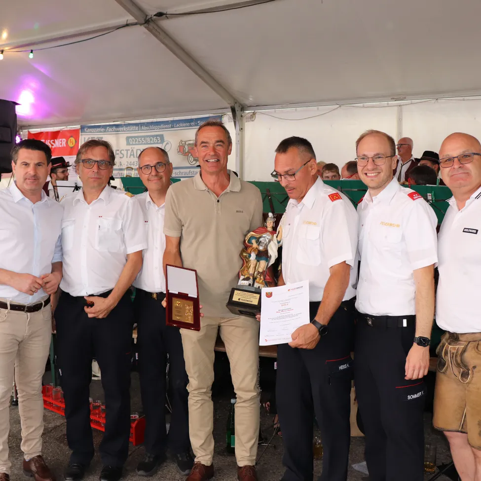 Sechs Männer in Uniformen und mit Brillen stehen in einem Zelt und lächeln für ein Foto. Ein Mann hält eine Trophäe und ein Zertifikat. Hinter ihnen ist ein Banner und eine Menschenmenge zu sehen.