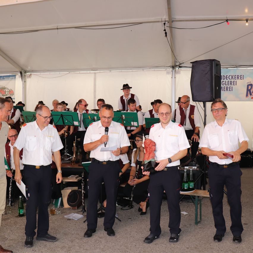 Eine Gruppe von Männern in weißen Uniformen steht in einer Reihe, einer hält ein Mikrofon und ein anderer ein Pokal. Hinter ihnen spielt eine Band, und ein Publikum schaut zu.