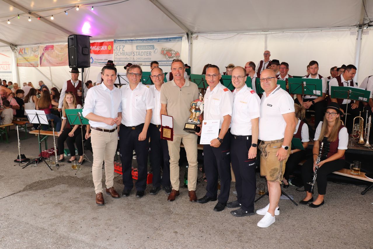 Eine Gruppe von Männern in Uniformen und Freizeitkleidung steht unter einem Zelt, hält einen Pokal und ein Zertifikat. Sie lächeln und posieren für ein Foto. Dahinter ist ein Banner mit Text und Logo zu sehen.